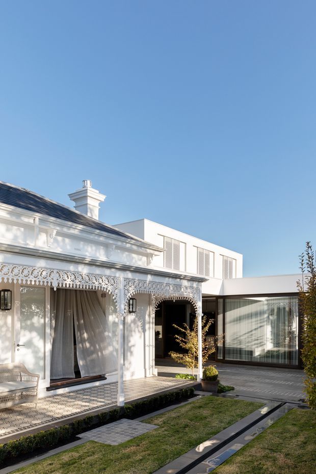 A bright courtyard exterior showing a restored ornate white heritage-style frontage with elaborate lacework veranda detailing, decorative columns, a tiled porch and chimney, connected to a clean-lined contemporary white and glazed extension. The landscaped courtyard includes clipped hedging, lawn, geometric paving and a narrow linear water feature.