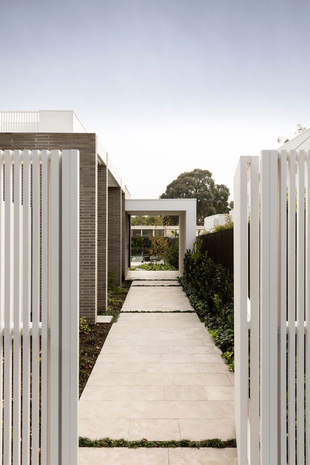 A narrow landscaped pedestrian passage between contemporary residential structures, viewed through open white vertical-slat gates. The walkway is paved with large pale stone or concrete slabs separated by planted joints, with clipped greenery along the sides. Dark textured brick walls, white rendered frames, and a rectangular portal structure lead toward a small courtyard or garden beyond. The composition is clean, geometric, and minimalist, with soft overcast lighting.