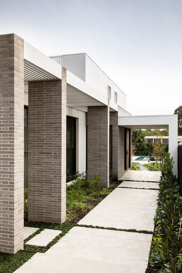 A contemporary residential exterior viewed along a landscaped pedestrian path. The building features pale grey-brown brick columns and walls, crisp white rendered volumes, deep roof overhangs with linear soffit detailing, narrow dark-framed windows, and a flat-roofed connecting volume. Large pale concrete or stone paving slabs form the walkway, with planted garden beds and a glimpse of a swimming pool beyond.