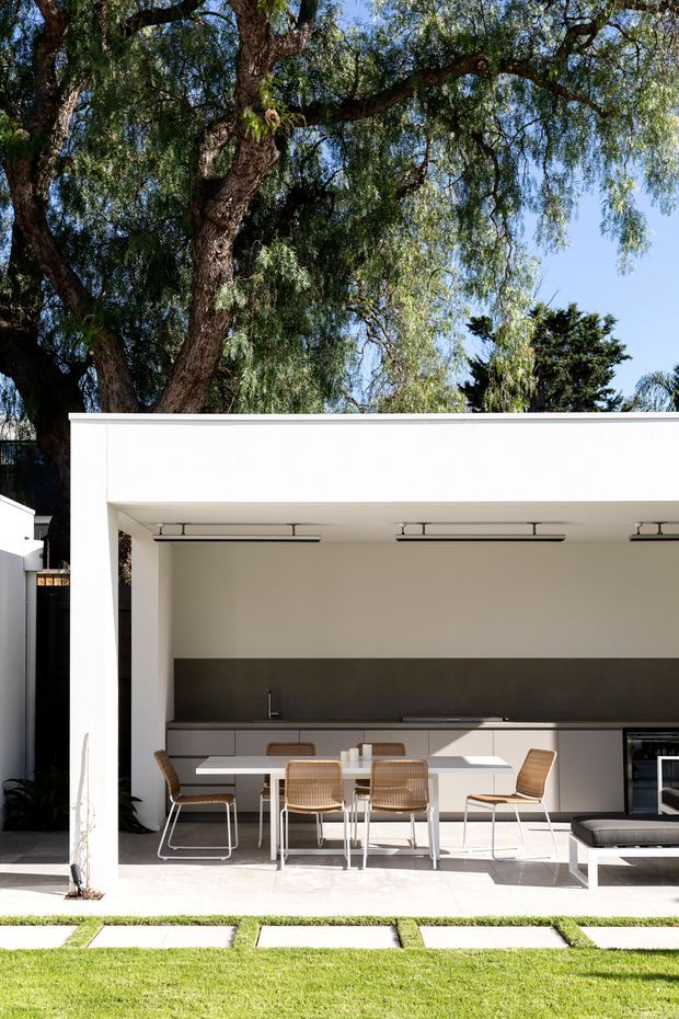 A bright, modern outdoor entertaining area with a white flat-roofed pavilion or open extension. The space contains a long outdoor kitchen with dark backsplash and built-in cabinetry, a white dining table surrounded by woven and metal-framed chairs, linear ceiling lights, and a low outdoor lounge seat. Large mature trees frame the structure, with a manicured lawn and rectangular stepping stones in the foreground.