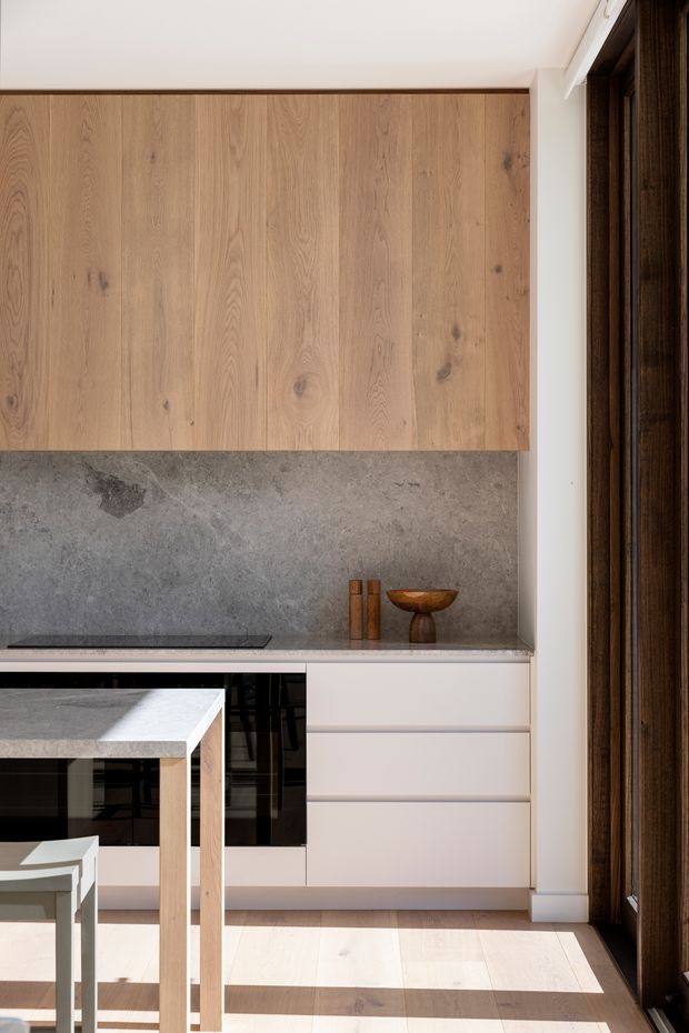 Bright contemporary kitchen with full-height natural timber cabinetry, a large grey stone splashback and benchtop, pale cabinetry, integrated dark appliances, and a compact dining table beside full-height dark timber-framed glazing. Strong sunlight creates long shadows across the pale timber floor.