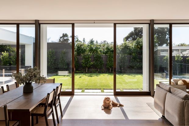 A bright, open-plan living and dining area with full-height glazed sliding doors opening onto a private, landscaped lawn. The interior features a long timber dining table with dark chairs, a neutral upholstered sofa, pale flooring, and generous natural light. Slim dark timber or metal-framed glazing creates strong views and shadows across the room.