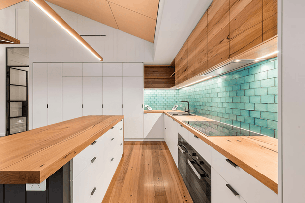 Bright contemporary galley kitchen with a large timber-topped island, white flat-panel cabinetry, warm timber flooring, extensive timber overhead joinery, and a turquoise glazed brick tile splashback. Integrated linear lighting runs beneath the upper cabinets and along the angled ceiling, creating a clean yet warm atmosphere.