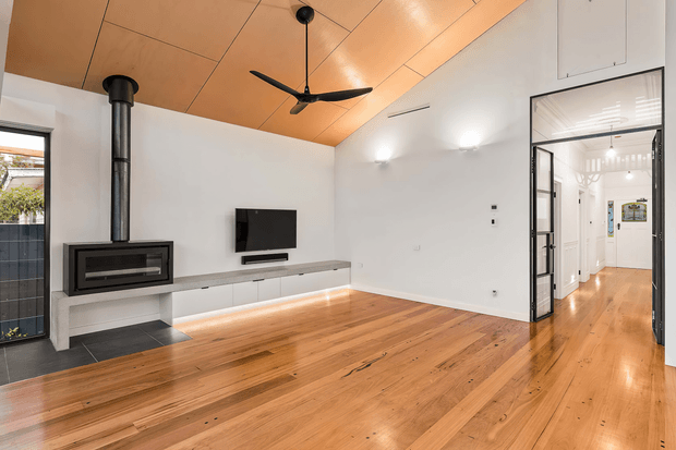 Bright contemporary living room with polished timber flooring, a warm timber panelled ceiling, white walls, a black wood-burning fireplace with exposed flue, wall-mounted television, ceiling fan, and built-in low cabinetry. Glazed black-framed doors open to a white hallway beyond.