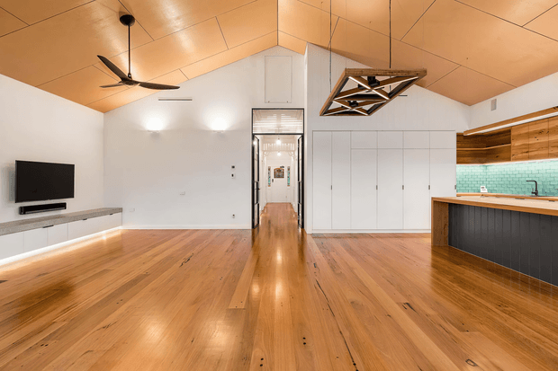 Open-plan contemporary living area with warm timber flooring and a striking timber-paneled ceiling. The room includes a wall-mounted television with low built-in joinery, a ceiling fan, central circulation toward a hallway, and a compact kitchen at right with a timber benchtop, dark island cabinetry, and turquoise tiled splashback. White built-in storage provides a clean backdrop, while a geometric timber-and-metal pendant adds a feature element.
