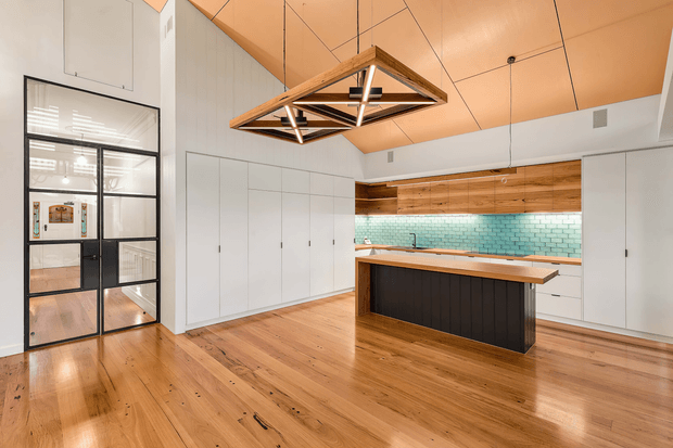 A spacious contemporary kitchen with warm timber flooring and a timber-lined ceiling, white full-height cabinetry, a central island with a dark vertical-panelled base and timber benchtop, and a turquoise glazed-tile splashback. A black-framed glazed door and partitions sit to the left, while a geometric timber-and-black pendant light hangs above the island.