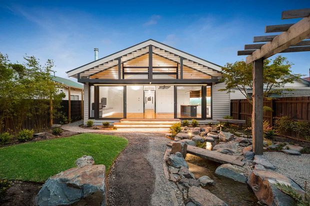 Single-storey gabled house viewed from the landscaped rear garden. The facade combines horizontal light-coloured cladding, extensive glazed sliding doors, and a timber-framed gable with exposed rafters and screened ventilation detailing. A paved and timber step terrace opens to a structured garden with lawn, rocks, gravel, planting, and a timber pergola or outdoor structure.