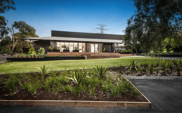 A single-storey contemporary building set within a landscaped suburban property. The low, elongated facade has a broad flat roof, dark horizontal cladding, extensive floor-to-ceiling glazing, and a covered entry or terrace. Timber decking and raised garden beds lead across a manicured lawn, with structured drought-tolerant planting in the foreground and mature trees around the site.