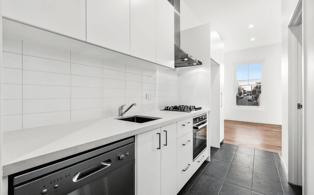 A bright, compact contemporary kitchen viewed along a linear run of white cabinetry and benchtop. The kitchen features a white tiled splashback, integrated stainless-steel dishwasher, black inset sink, gas cooktop, stainless-steel rangehood and built-in oven. Dark grey floor tiles contrast with the white joinery, while a timber-floored adjoining area is visible beyond the kitchen.