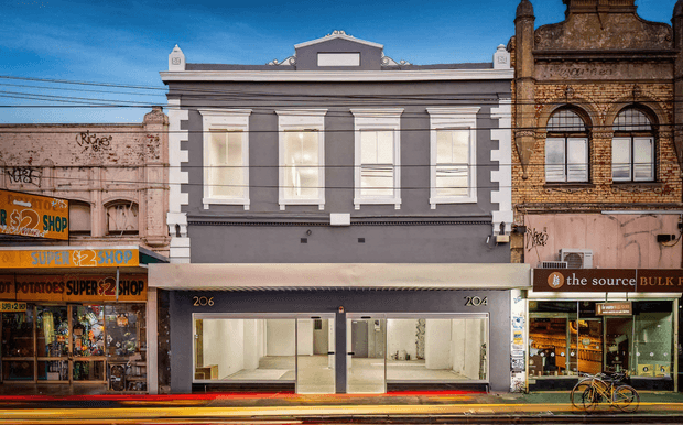 Street-facing view of a renovated multi-storey commercial building with a symmetrical grey and white classical facade, tall upper-level windows, ground-floor retail glazing, awnings, and neighbouring heritage shopfronts in an urban streetscape.