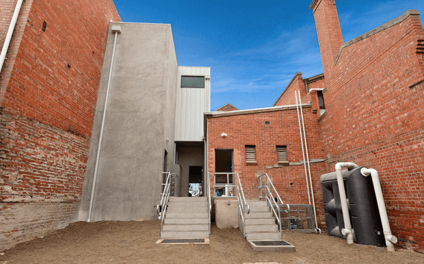 Exterior view of a compact contemporary addition or service area enclosed by tall red-brick walls. The scene features a raw concrete tower-like volume, corrugated metal cladding, brick chimney forms, external stairs with stainless-steel handrails, and large exposed black-and-white pipes or tanks. The image has a utilitarian, industrial character under a clear blue sky.