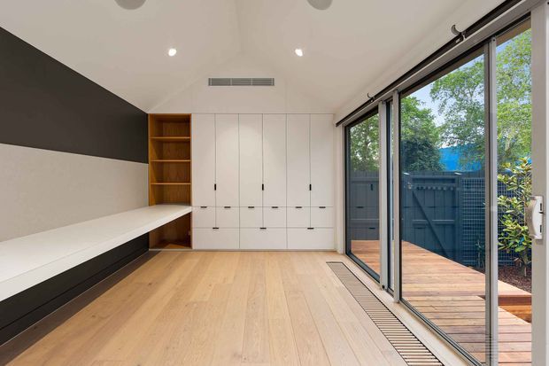 Bright, compact contemporary room with pale timber flooring, a built-in white storage wall and open shelving, and a long built-in bench or daybed along a two-tone feature wall. Full-height sliding glass doors open directly to a timber deck and enclosed garden courtyard, bringing in abundant natural light.