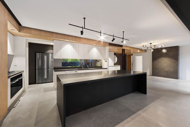 A spacious contemporary open-plan kitchen with a large black island, white handleless cabinetry, integrated appliances, dark stone-look splashback and wall, warm timber ceiling and wall detailing, and polished concrete flooring. Linear track lighting and sculptural pendant fixtures illuminate the kitchen and adjacent open living area.