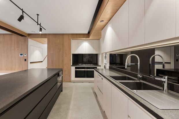 Contemporary galley-style kitchen with a large dark island, white handleless cabinetry, integrated appliances, double sinks and metallic fixtures. Warm timber wall and ceiling accents contrast with the white cabinetry, dark reflective splashback and polished grey benchtops. Black track lighting adds a graphic detail to the otherwise clean, refined interior.