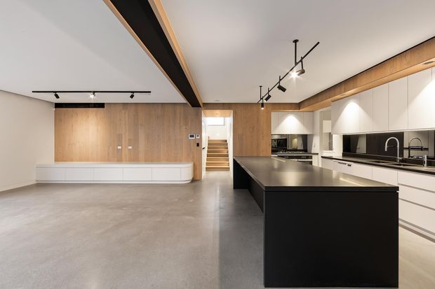 A spacious contemporary open-plan kitchen and living area with a large dark island, white flat-panel cabinetry, black splashback, timber wall panelling, and polished concrete flooring. Linear and track lighting complement the exposed dark ceiling beam, while stairs lead to an upper level at the rear.