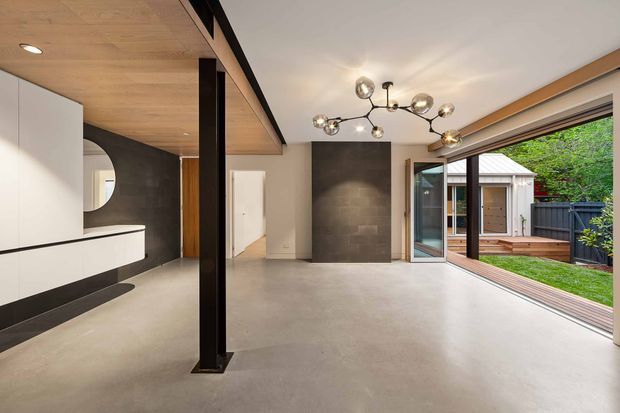 Open-plan contemporary interior with a polished concrete floor, dark stone feature wall, timber-lined ceiling and built-in white cabinetry. A black structural post divides the space, while large sliding glass doors open to a timber deck and landscaped courtyard. A sculptural multi-globe pendant provides a prominent lighting feature.