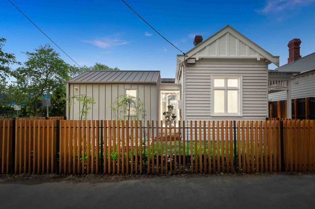 Street-facing view of a compact residential property with a restored-looking weatherboard cottage facade, decorative gable trim, brick chimney and corrugated metal-roofed rear extension. A timber picket fence and small landscaped garden sit in front of the home.