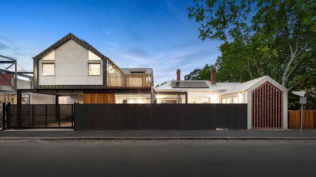 Street-facing dusk view of a contemporary multi-level residential property combining a dark-clad upper gabled volume with a lighter traditional dwelling behind a high front fence. The facade includes timber-look vertical screening, balconies, large windows, a secure driveway or garage entry, and a separate pitched-roof structure with a strong timber slat gable. Mature trees frame the suburban setting.
