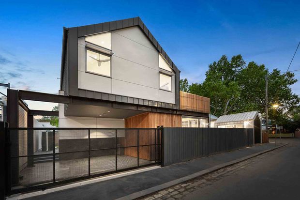 Street-facing view of a contemporary multi-storey urban residence with a sharply pitched gable roof, dark metal cladding and trim, light rendered wall panels, timber screening, and large upper-level windows. A covered carport or entry canopy sits behind a black metal fence, with neighbouring landscaping and a small glazed structure visible to the side.
