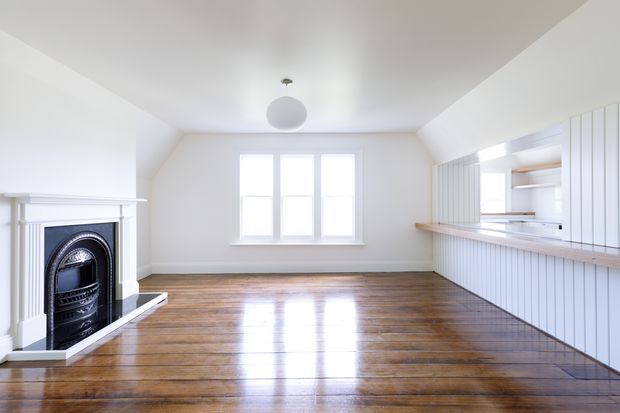 Bright, largely empty open-plan interior with polished timber flooring, a traditional white fireplace at left, and a white vertical-panelled kitchen or bar joinery area with a timber counter along the right wall. A central multi-pane window provides daylight, while a simple spherical pendant light hangs from the flat white ceiling. The room has a clean, airy transitional character with traditional architectural details.