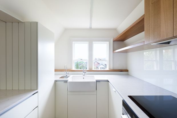 Bright, compact contemporary kitchen with a white L-shaped worktop, integrated cabinetry, a large white farmhouse-style sink, timber open shelving and upper cabinetry, and a black induction cooktop. A window provides natural light at the rear, while a tall bank of pale vertical-panel cabinetry occupies the left side.