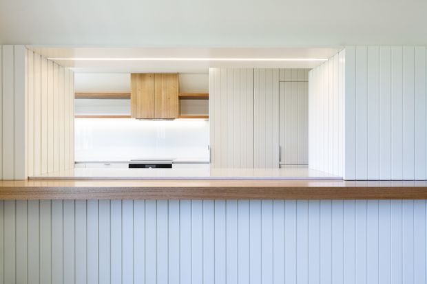 A bright, minimalist kitchen viewed across a substantial timber-topped island or peninsula. The cabinetry and surrounding walls feature vertical white painted panelling, with open timber shelving, a simple timber wall cabinet, concealed appliances, and integrated linear lighting. The composition is clean and highly symmetrical, with pale finishes and warm wood accents.