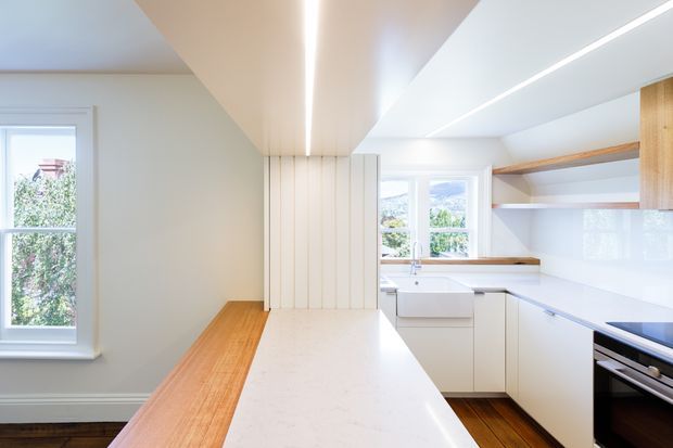 Bright contemporary kitchen with a long white stone island, integrated white cabinetry, timber accents, timber flooring, and linear recessed ceiling lighting. A stainless-steel oven is visible beneath the benchtop, with large windows bringing in natural light and views of greenery.