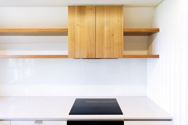 A bright minimalist kitchen vignette with a white stone benchtop, black induction cooktop, glossy white splashback, open timber shelves, and a central wall-mounted timber cabinet. Vertical white wall panelling is visible at the right edge.