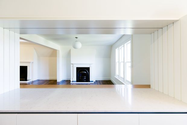 Bright white kitchen interior viewed from behind a large pale stone benchtop, looking toward an adjacent open living area with a traditional fireplace. The kitchen has white fixed cabinetry, vertical wall panelling, a reflective light-toned ceiling or overhead surface, and large multi-pane windows. A simple white pendant light and dark timber flooring are visible in the adjoining space.