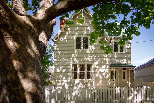 A two-storey heritage-style timber-clad house viewed from the street, partially framed by a large mature tree. The pale weatherboard facade features decorative gable detailing, ornate bargeboards, sash-style windows, a brick chimney, and a covered entry porch behind a white picket fence. Strong dappled sunlight creates a leafy, established residential setting.