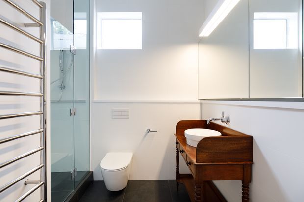 Bright contemporary bathroom with a frameless glass shower enclosure, wall-mounted mirrored cabinetry, a white toilet, dark tiled flooring, and a distinctive timber washstand with a white basin. A chrome heated towel rail occupies the left side, and concealed linear lighting runs beneath the upper cabinets.