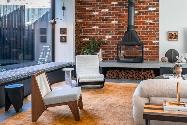 A sophisticated living room with an exposed red-brick feature wall, built-in black fireplace and firewood storage, expansive glazing, and a mix of sculptural contemporary and mid-century-inspired furniture. Warm neutral furnishings and a large tan rug create a refined, inviting atmosphere.
