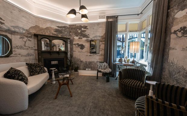 Elegant, richly layered living room with a curved cream sofa, striped velvet armchairs, ornate dark fireplace surround, patterned scenic wallcovering, full-height drapery, a bay window, and decorative ceiling detailing. The room has a refined, moody boutique-hotel character with warm accent lighting and classic architectural elements.