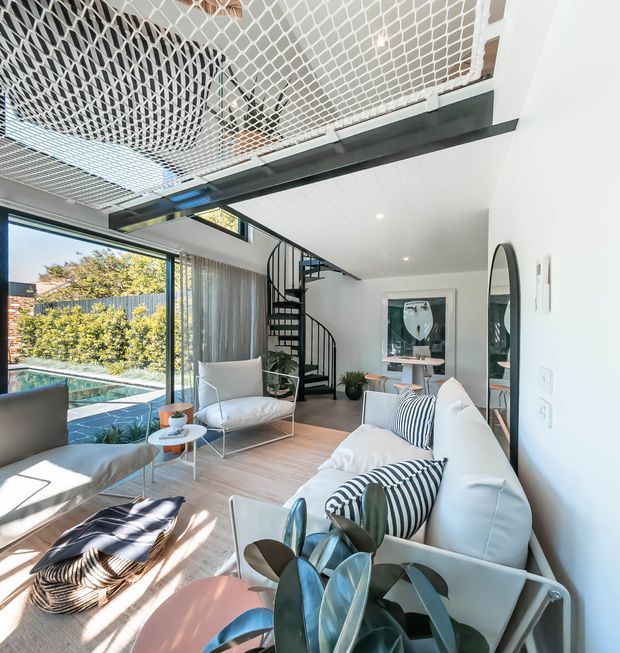 Bright contemporary living room with white sofas, striped cushions, a sculptural coffee table and indoor plants. Floor-to-ceiling glazing opens to an outdoor pool area, while a black spiral staircase leads to a mezzanine with a distinctive white safety net. Light timber flooring, white walls and black metal details create an airy, modern interior.
