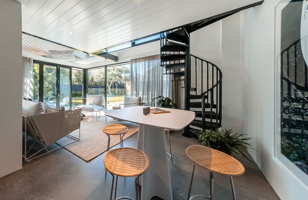 Bright contemporary open-plan living and dining area with a curved white dining table, woven bar stools, pale upholstered seating, concrete-look flooring, extensive glazing, and a striking black spiral staircase leading to a mezzanine. White panelled ceilings, indoor plants, and a light-filled garden outlook create an airy, modern interior.