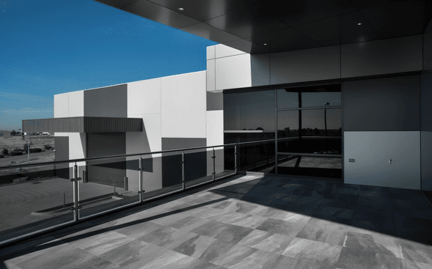 A modern commercial or institutional building exterior viewed from a covered terrace or elevated walkway. The scene features crisp white and grey geometric wall volumes, extensive dark glazing, a black soffit, metal-and-glass balustrades, and large-format grey paving. The architecture has a minimalist contemporary character with strong horizontal and cubic forms.
