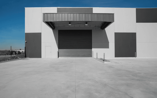 A stark, modern industrial building facade with a broad concrete forecourt, large dark roller door, projecting metal awning, and bold white and charcoal geometric wall panels. The image is presented in a cool, high-contrast monochrome-like palette with a small amount of blue sky visible.