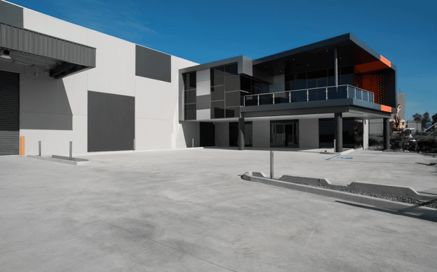Contemporary commercial or industrial building exterior with a broad concrete forecourt, large warehouse-style wall panels and roller door, and a two-storey dark-clad office or entry volume with balcony, glazing, and projecting flat roof. The composition is strongly geometric, with white and charcoal façades and a small orange accent panel.