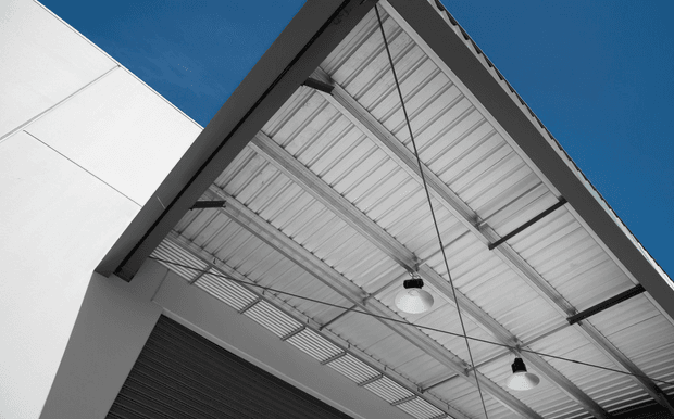 Upward-looking architectural view of a covered exterior canopy or verandah with a pitched corrugated metal roof, exposed steel framing and rafters, suspended pendant lights, and clean white wall surfaces against a blue sky.