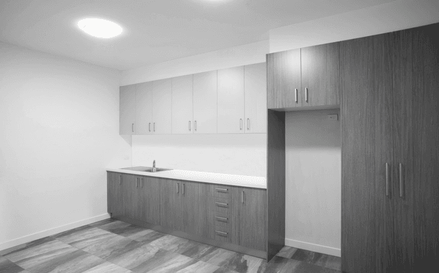 A compact, modern kitchen with extensive grey wood-look cabinetry, white overhead cupboards, a pale benchtop and simple integrated sink. Full-height pantry-style storage is visible on the right, with grey timber-look flooring and recessed ceiling lighting. The room is empty and functional, with a neutral monochromatic palette.
