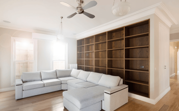 Bright, refined living room featuring a large pale sectional sofa with ottoman seating, a full-height dark timber built-in shelving wall, light timber flooring, white detailed cornices and wall panelling, pendant lights, and a ceiling fan. A hallway is visible through the opening at right.