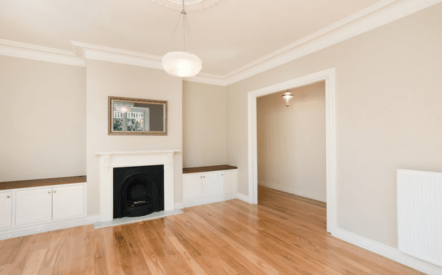 Bright, empty traditional-style living room with polished timber flooring, beige walls, white decorative cornices and trim, built-in low cabinetry with dark benchtops, and a white mantel surrounding a black fireplace. A wide trimmed doorway opens to an adjoining hall or room, with pendant lighting and a wall radiator visible.