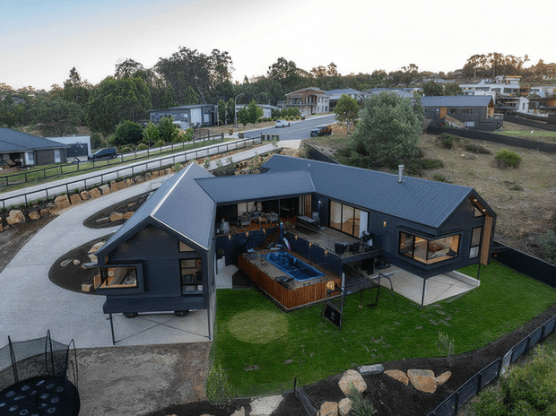 Aerial view of a contemporary single-storey dark-clad house on a landscaped suburban or semi-rural site. The home has several intersecting pitched roof volumes, extensive glazing, an outdoor entertaining area with a spa or plunge pool, and a manicured lawn enclosed by dark fencing. Curved concrete driveways and neighbouring modern homes are visible around the property.