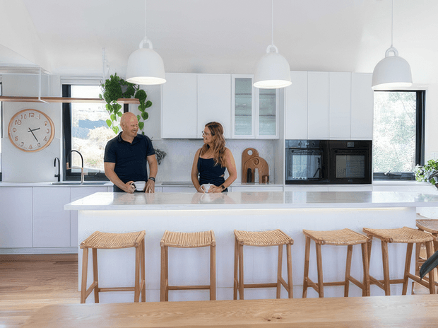 Bright, spacious contemporary kitchen with extensive white cabinetry, a large white island with integrated seating, black-framed windows, timber flooring, and three white pendant lights. Natural timber bar stools and a timber dining table add warmth, while integrated black appliances and a stone-look benchtop provide contrast. Two people are standing at the island, with indoor greenery and simple timber accessories visible.