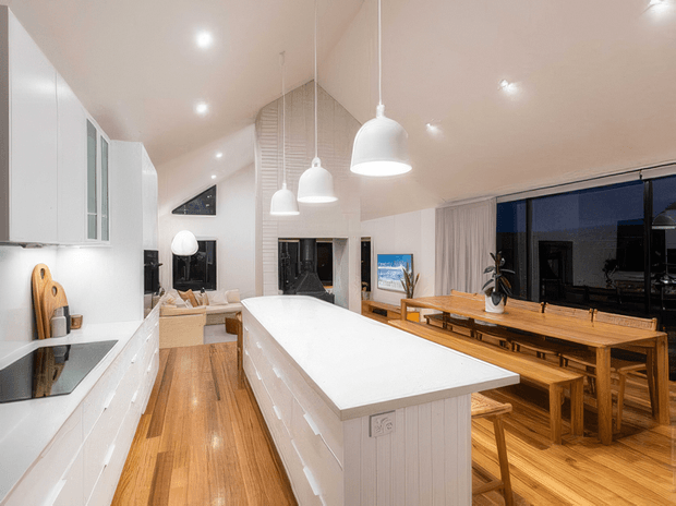 Bright open-plan kitchen and dining area with a large white island, white cabinetry, timber flooring, pendant lights, and a long timber dining table beside dark-framed glazing. The space has a warm, clean contemporary coastal feel with a pitched ceiling and vertical timber wall detailing.