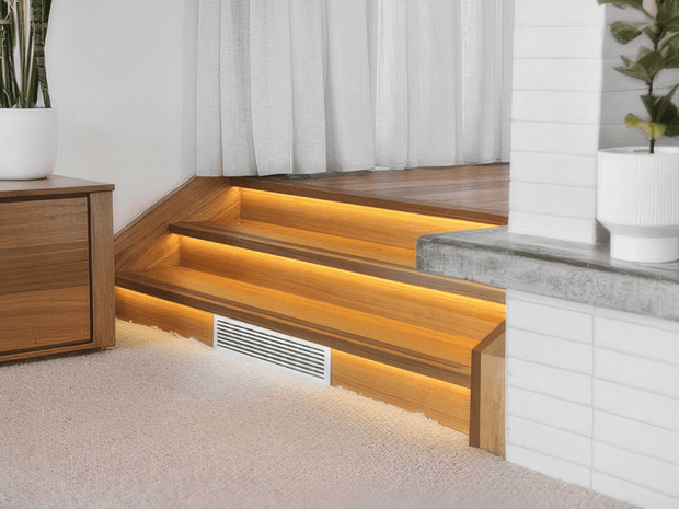Interior view of a warm, contemporary split-level transition with three illuminated timber-clad steps leading to a raised platform. A linear air vent is integrated into the lower riser, while a white tiled fireplace or partition with a grey concrete ledge frames the right side. Sheer white curtains, pale carpet, timber joinery, and potted plants complete the calm residential setting.