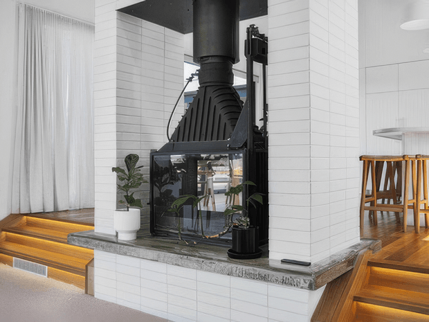 Bright contemporary interior centered on a large black freestanding fireplace set within a white tiled masonry surround and raised hearth. The fireplace has a dark metal flue and visible firebox, with plants placed on the hearth. White vertical curtains, warm timber flooring, and stepped floor levels frame the space; a kitchen or dining area with timber stools is visible to the right.