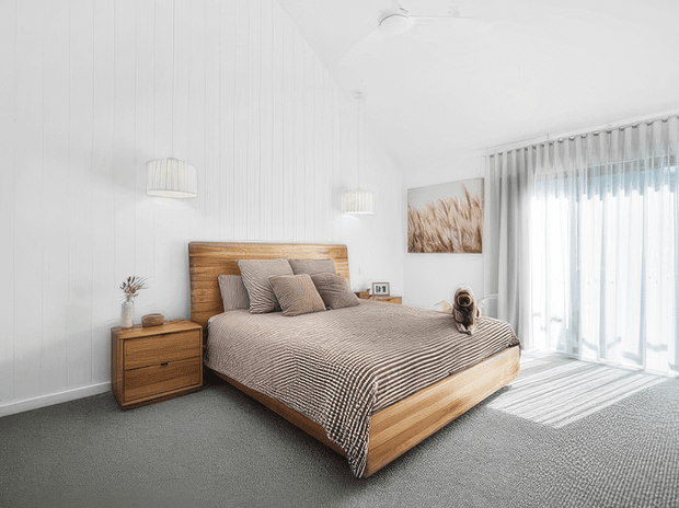 Bright, calm bedroom with a low-profile timber bed and matching timber bedside table, layered neutral striped bedding, grey carpet, white vertically panelled walls, pendant lights, and full-height sheer curtains opening to a sunlit exterior.