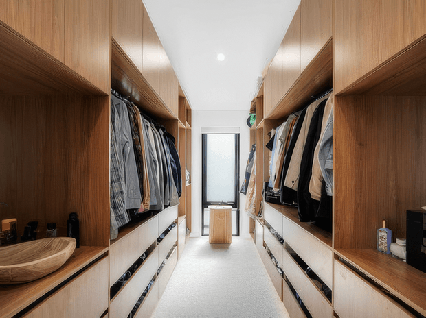 A bright, narrow walk-in wardrobe lined with symmetrical warm timber cabinetry, open hanging rails, and white drawer units on both sides. A small central timber island or bench sits in the walkway, with a frosted-glass door or window at the far end. Built-in shelving also displays a timber basin and small accessories.