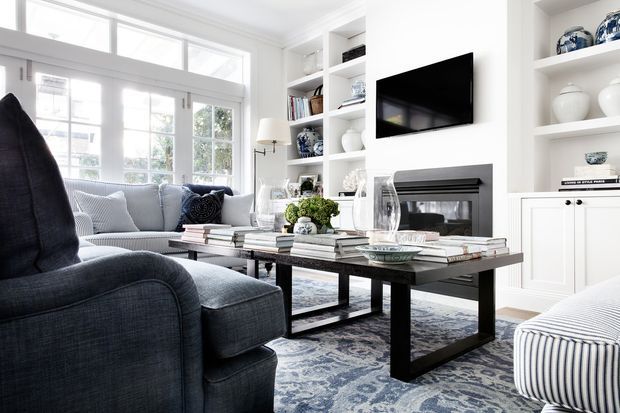 Bright, refined living room with large white-framed windows, built-in white shelving and cabinetry, a wall-mounted television, and a dark fireplace surround. The room features deep blue-grey upholstered seating, a black coffee table layered with books and decor, patterned rug, and blue-and-white accessories.